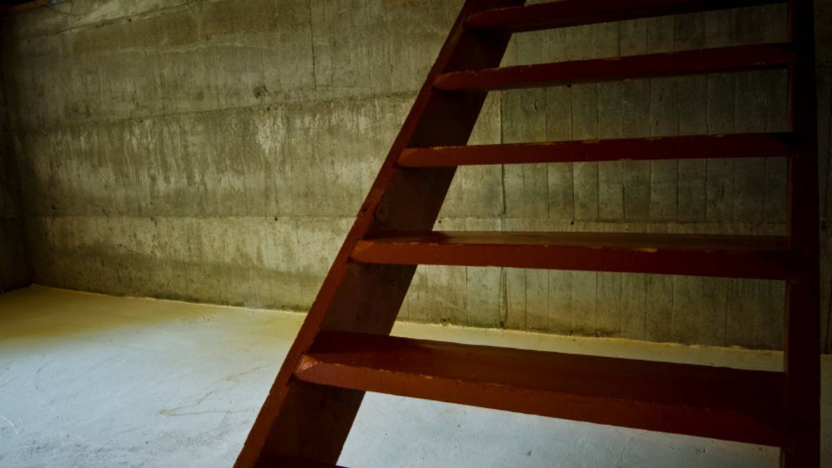 Een rode houten keldertrap staat voor een kale betonmuur waarop een horizontale vochtstreep loopt, met daaronder lichte uitslag langs de aansluiting met de grijze betonvloer.